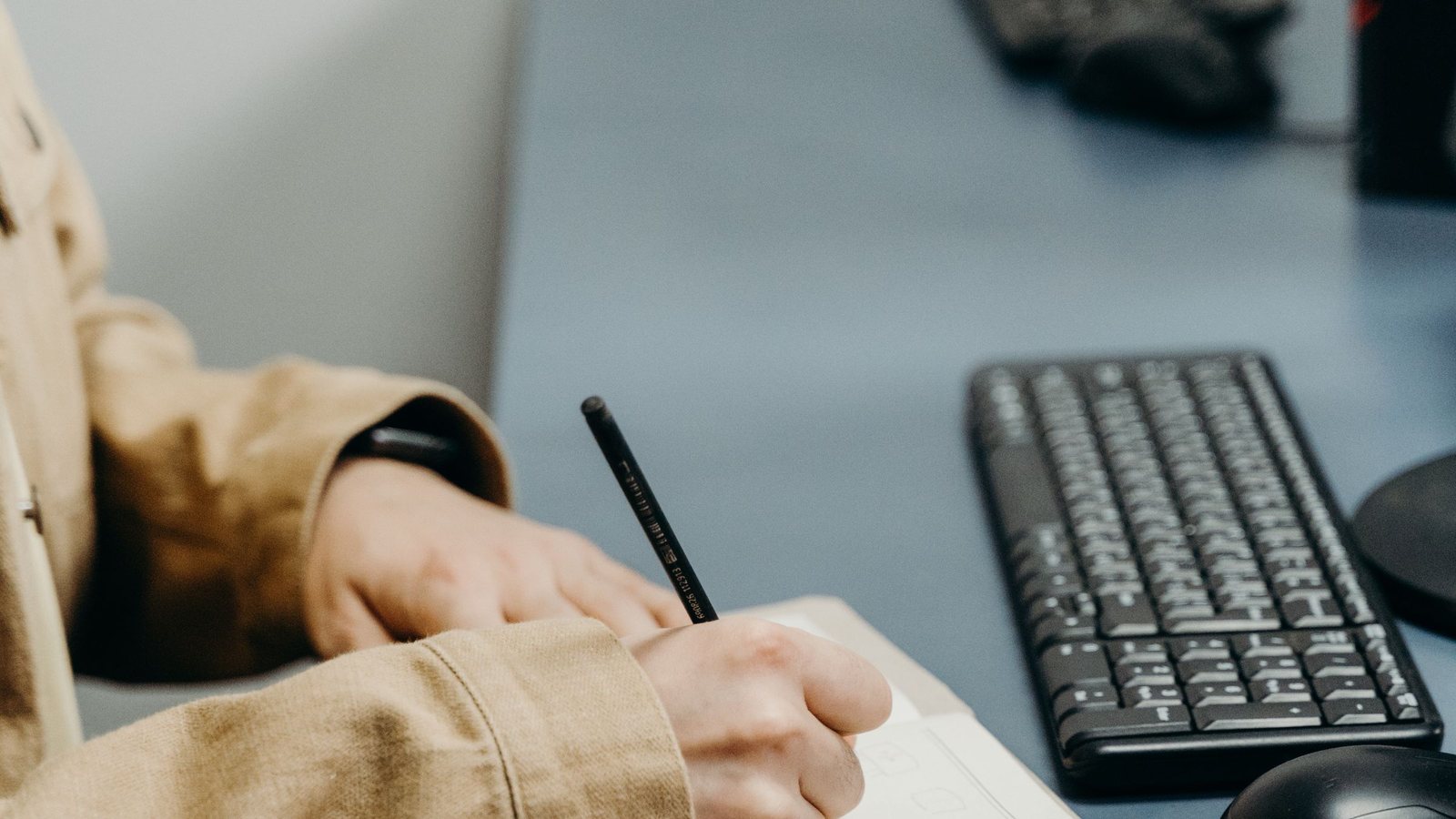 Person writing notes by hand at a desk beside a computer keyboard