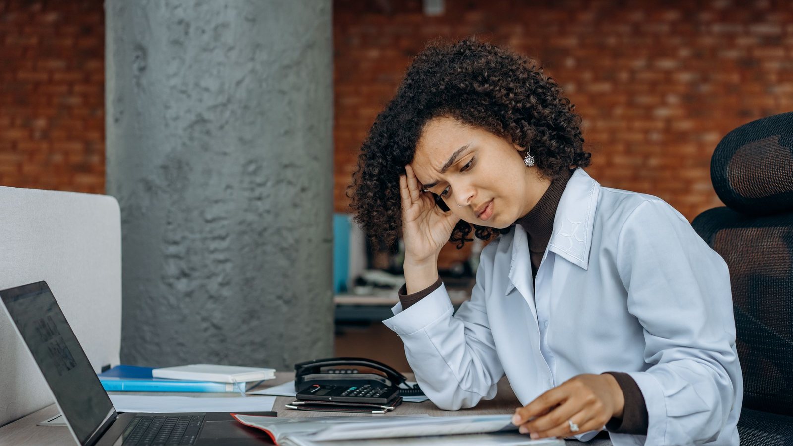 A stressed office worker holding her head while going through a binder of documents at her desk